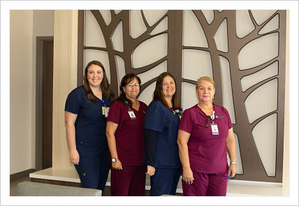This is a picture of four ladies in a hallway with tree art behind them. Two are wearing blue scrubs and two are wearing maroon scrubs.