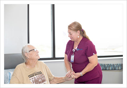 This is a picture of a nurse wearing maroon scrubs holding the hand of a male elderly patient.