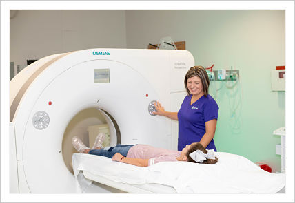 This is a picture of a MRI Machine with a little girl wearing a apink shirt with blue jeans and wearing a bow laying on a table about to go through the machine with a nurse standing by the machine fixing to turn the machine on.